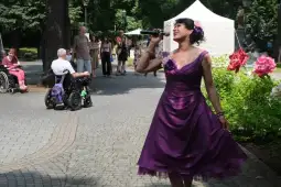 Kobieta w fioletowej sukience śpiewa, otoczona przez ludzi w parku.