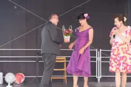 Mężczyzna wręcza kobietę w fioletowej sukni bukiet kwiatów na scenie. Obok stoi druga kobieta z mikrofonem.