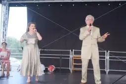Dwoje wykonawców śpiewa na scenie, obok siedzi kobieta.