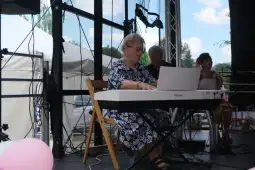 Starsza kobieta gra na pianinie na scenie, obok niej dwie inne osoby.
