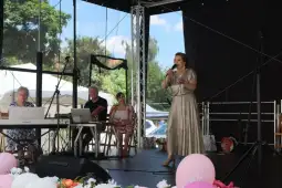 Kobieta w złotej sukni śpiewa na scenie, obok muzycy i publiczność.