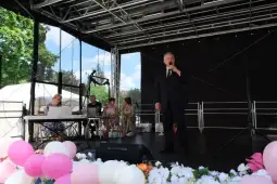 Mężczyzna w garniturze śpiewa na scenie, obok siedzą muzycy i widownia. Kolorowe balony w dolnej części.