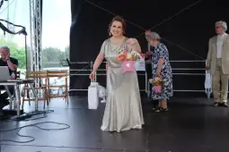 Kobieta w srebrnej sukni trzyma bukiet kwiatów na scenie, w tle inni uczestnicy wydarzenia.