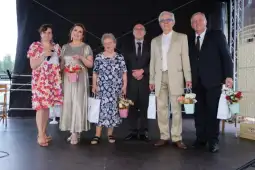 Grupa sześciu osób w eleganckich strojach, trzymających kwiaty i torby, na scenie.