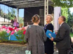Kobieta w okularach rozmawia z dwiema osobami, w tle występ artystyczny.