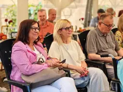 Grupa osób siedzących na krzesłach, uśmiechnięte kobiety w okularach, mężczyzna z laską.