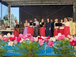 Grupa osób w strojach ludowych śpiewa na scenie, obok instrumenty muzyczne i dekoracje kwiatowe.