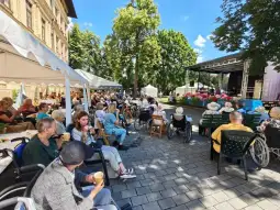 Wydarzenie plenerowe z uczestnikami siedzącymi na krzesłach i w wózkach inwalidzkich, scena w tle.
