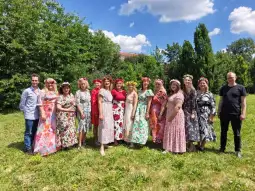 Grupa kobiet w kolorowych sukienkach z kwiatowymi wiankami na głowach, pozująca na tle zieleni.