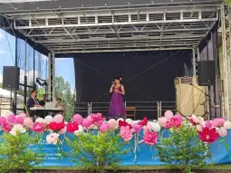 Kobieta w fioletowej sukience śpiewa na scenie, obok muzycy przy fortepianie. Dekoracje z kwiatów i balonów.