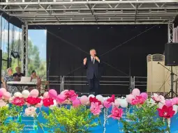 Mężczyzna w garniturze przemawia na scenie, w tle dekoracje z kwiatów i balonów.