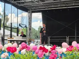 Mężczyzna w garniturze przemawia na scenie, obok pianista, w tle kwiaty i balony.