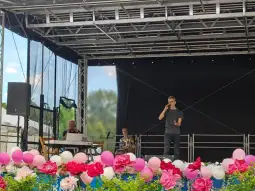 Mężczyzna śpiewa na scenie, obok dwie osoby przy instrumentach, w tle dekoracje z kwiatów.
