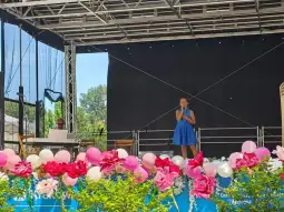 Dziewczynka w niebieskiej sukience śpiewa na scenie, otoczona kwiatami i balonami.