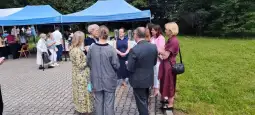 Grupa ludzi rozmawia w plenerze, obok namioty i zieleń.