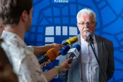 Mężczyzna w okularach przemawia do mikrofonu, otoczony przez dziennikarzy z mikrofonami.