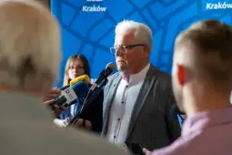 Mężczyzna w okularach przemawia do mikrofonu, w tle dziennikarze.