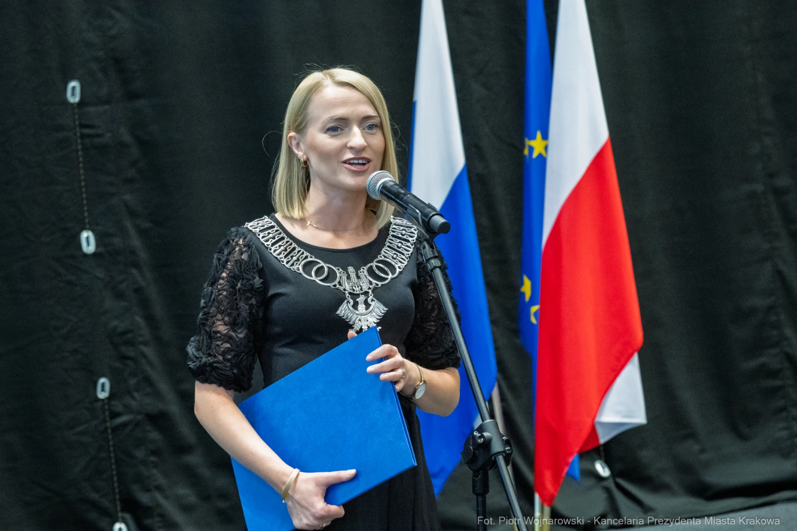 Kobieta przemawia do mikrofonu, trzymając niebieski dokument, w tle flagi Polski i Unii Europejskiej.  Autor: Piotr Wojnarowski
