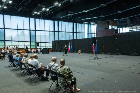 Spotkanie w sali z mówcą, publiczność siedząca na widowni, flagi Polski w tle.