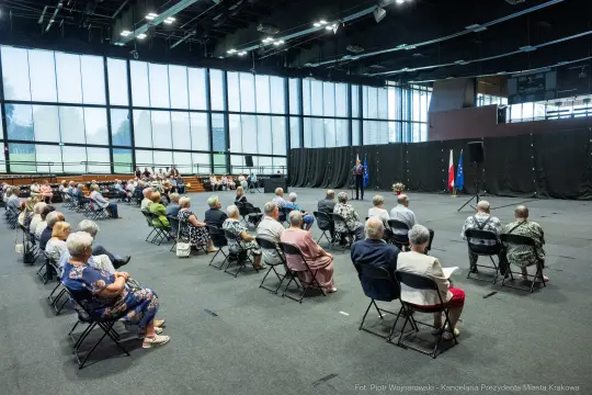 Spotkanie osób starszych w sali, z mówcą na podium i flagami w tle.