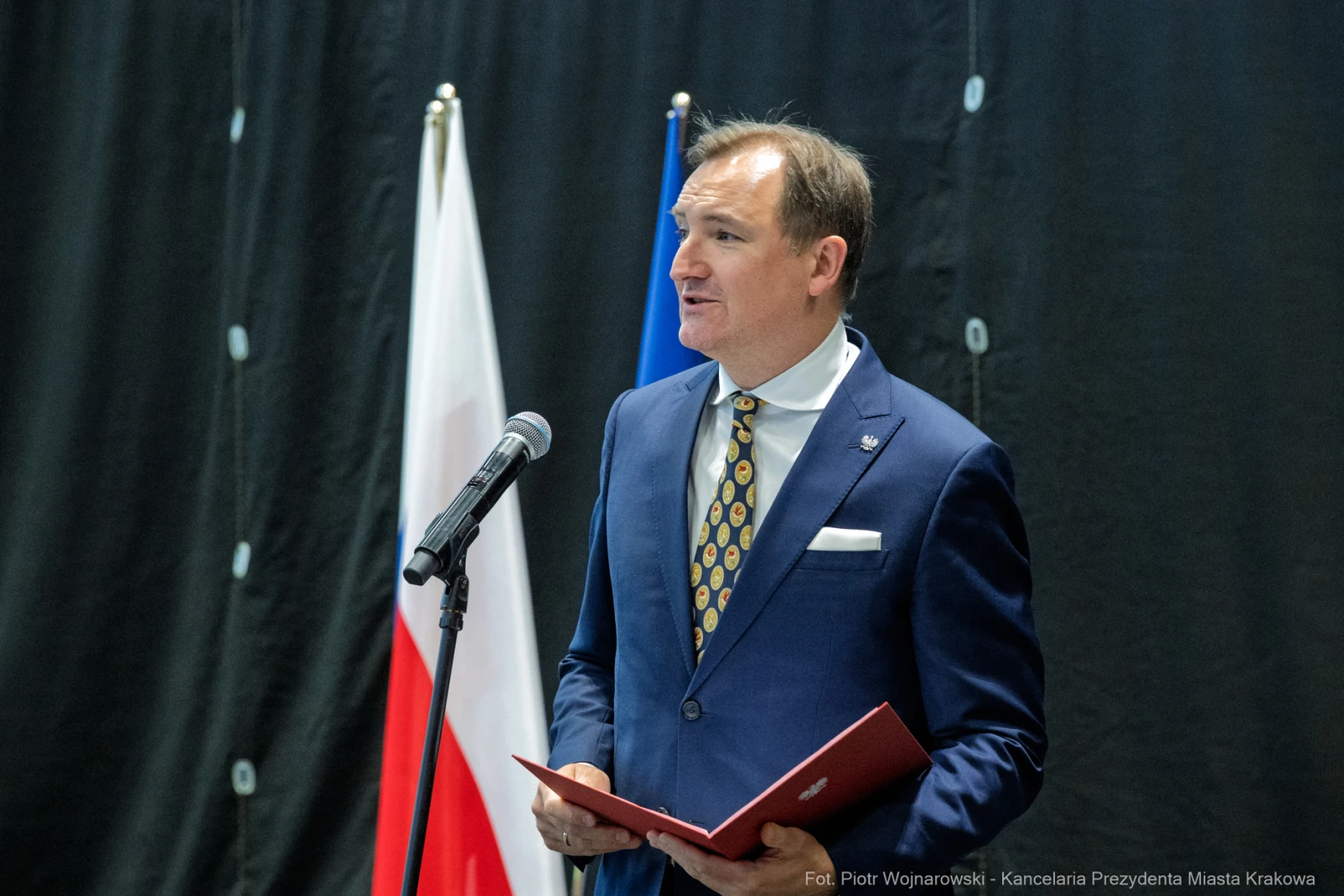 Mężczyzna w garniturze przemawia przy mikrofonie, obok flagi Polski i Unii Europejskiej.  Autor: Piotr Wojnarowski