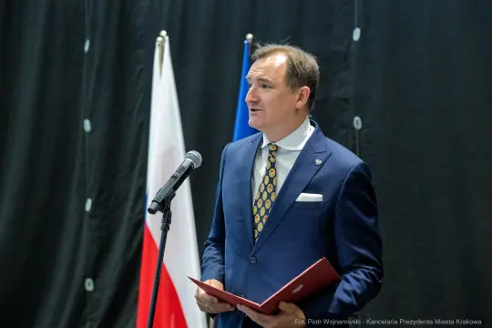 Mężczyzna w garniturze przemawia przy mikrofonie, obok flagi Polski i Unii Europejskiej.