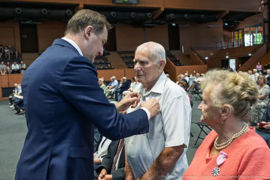 Uroczystość wręczenia odznaczeń, mężczyzna przypina medal starszemu mężczyźnie. Obok stoi kobieta.