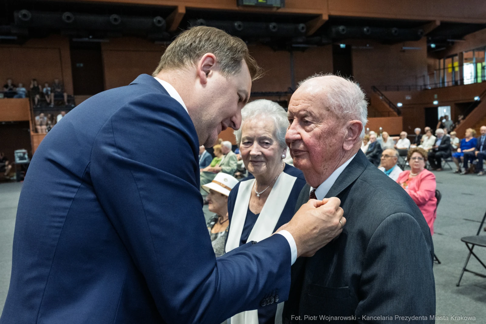 Mężczyzna przypina odznaczenie starszemu mężczyźnie w obecności kobiety. W tle siedzą goście.  Autor: Piotr Wojnarowski