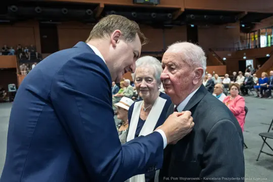 Mężczyzna przypina odznaczenie starszemu mężczyźnie w obecności kobiety. W tle siedzą goście.