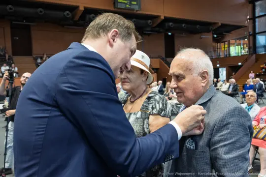 Mężczyzna przypina odznaczenie starszemu mężczyźnie w towarzystwie kobiety.