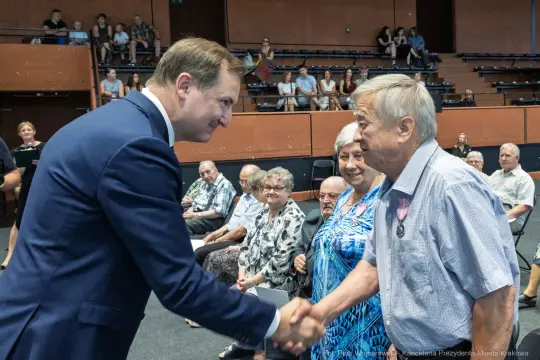 Uścisk dłoni między mężczyzną w garniturze a starszym mężczyzną, w tle siedzący publiczność.