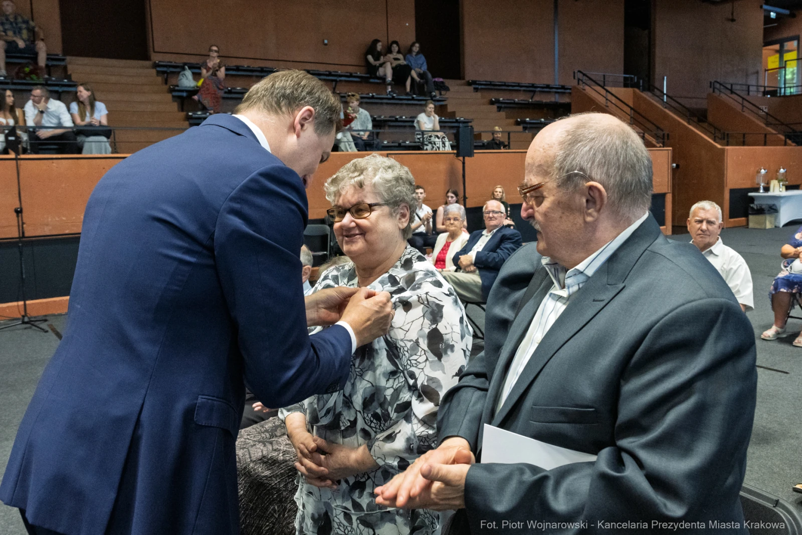 Wręczenie odznaczenia kobiecie przez mężczyznę w formalnym otoczeniu. W tle zgromadzeni goście.  Autor: Piotr Wojnarowski