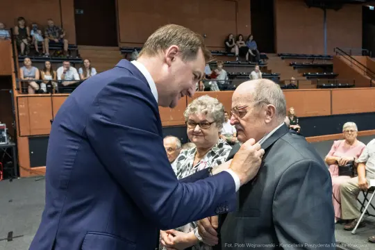 Uroczystość wręczenia odznaczeń, mężczyzna przypina medal starszemu mężczyźnie.