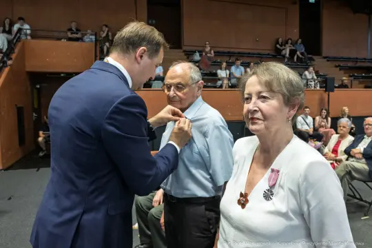 Wręczenie odznaczenia mężczyźnie przez urzędnika w obecności kobiety i innych gości.