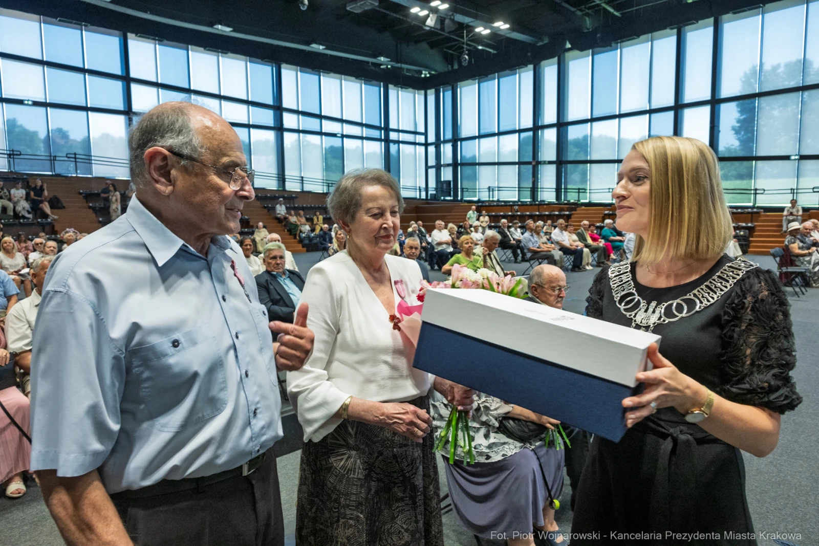 Wręczenie prezentu starszej parze przez kobietę w eleganckim stroju. W tle zgromadzenie osób.  Autor: Piotr Wojnarowski