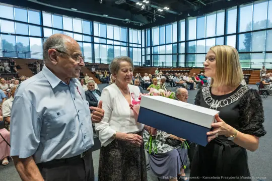 Wręczenie prezentu starszej parze przez kobietę w eleganckim stroju. W tle zgromadzenie osób.