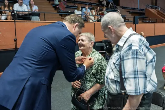Wręczenie odznaczenia starszej kobiecie przez mężczyznę w garniturze, obok stoi starszy mężczyzna.