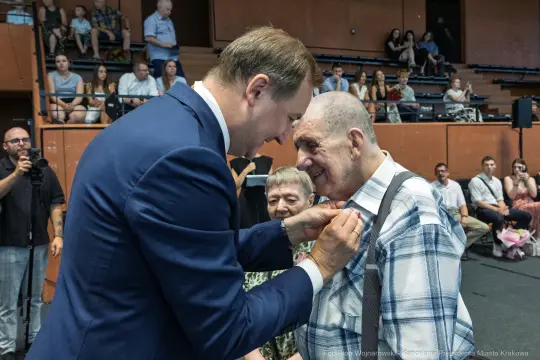 Uroczystość wręczenia odznaczenia starszemu mężczyźnie przez uśmiechniętego mężczyznę w garniturze.