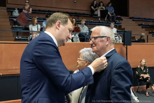 Mężczyzna przypina odznaczenie innemu mężczyźnie w obecności kobiety.