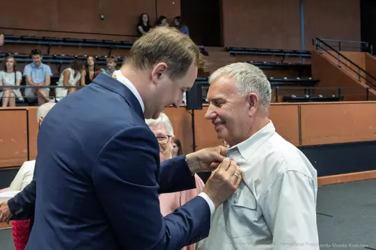 Wręczenie odznaczenia mężczyźnie przez innego mężczyznę w formalnej atmosferze.