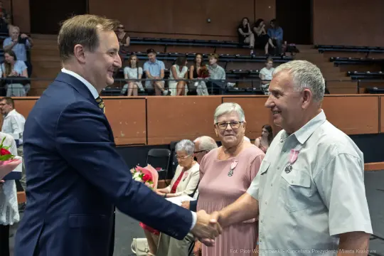 Uroczystość wręczenia odznaczeń, mężczyzna podaje rękę innemu mężczyźnie. W tle widoczna publiczność.