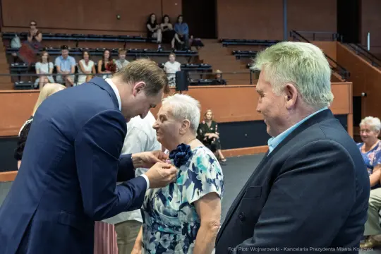 Uroczystość wręczenia odznaczeń, uśmiechnięte twarze, eleganckie stroje.