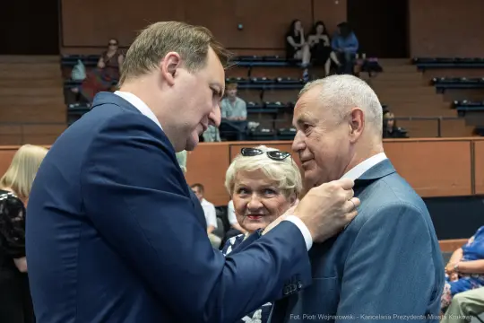 Mężczyzna przypina odznaczenie do marynarki innego mężczyzny, w tle kobieta obserwuje.
