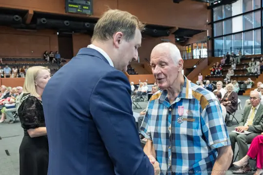 Uroczystość wręczenia odznaczeń, mężczyzna w koszuli w kratę rozmawia z innym mężczyzną.
