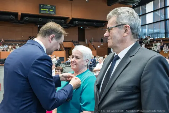 Wręczenie odznaczenia przez mężczyznę starszej kobiecie w towarzystwie drugiego mężczyzny.
