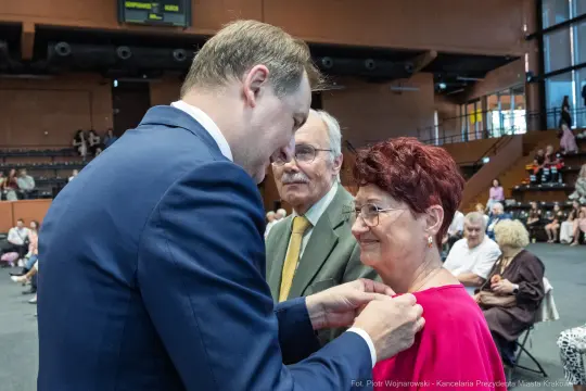 Wręczenie odznaczenia kobiecie przez mężczyznę, w tle starsza para.