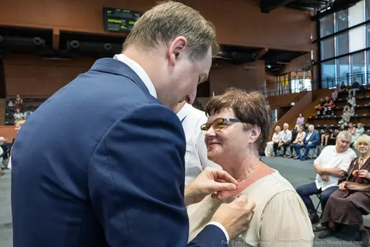 Uroczystość wręczenia odznaczenia przez mężczyznę kobiecie w eleganckim stroju.