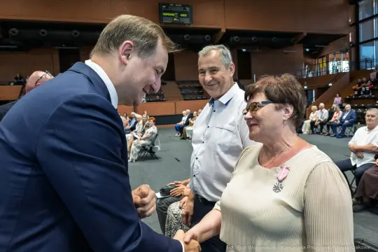 Spotkanie dwóch osób, uściski dłoni, uśmiechy, sala wypełniona ludźmi.