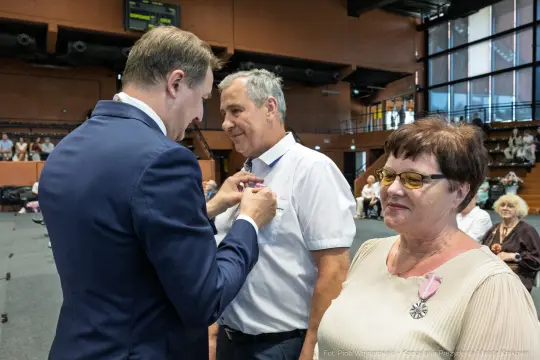 Wręczenie odznaczenia mężczyźnie przez ubranego w garnitur mężczyznę, obok stoi kobieta.