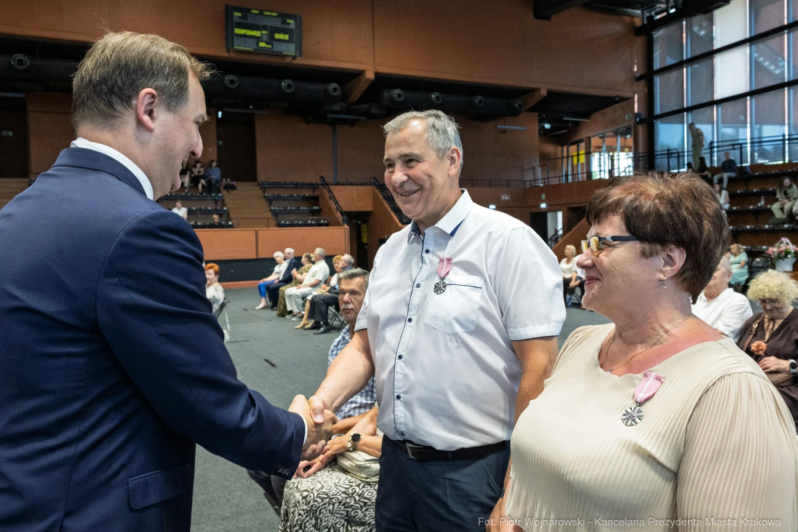 Spotkanie trzech osób, uściski dłoni i uśmiechy w tle sala z publicznością.  Autor: Piotr Wojnarowski
